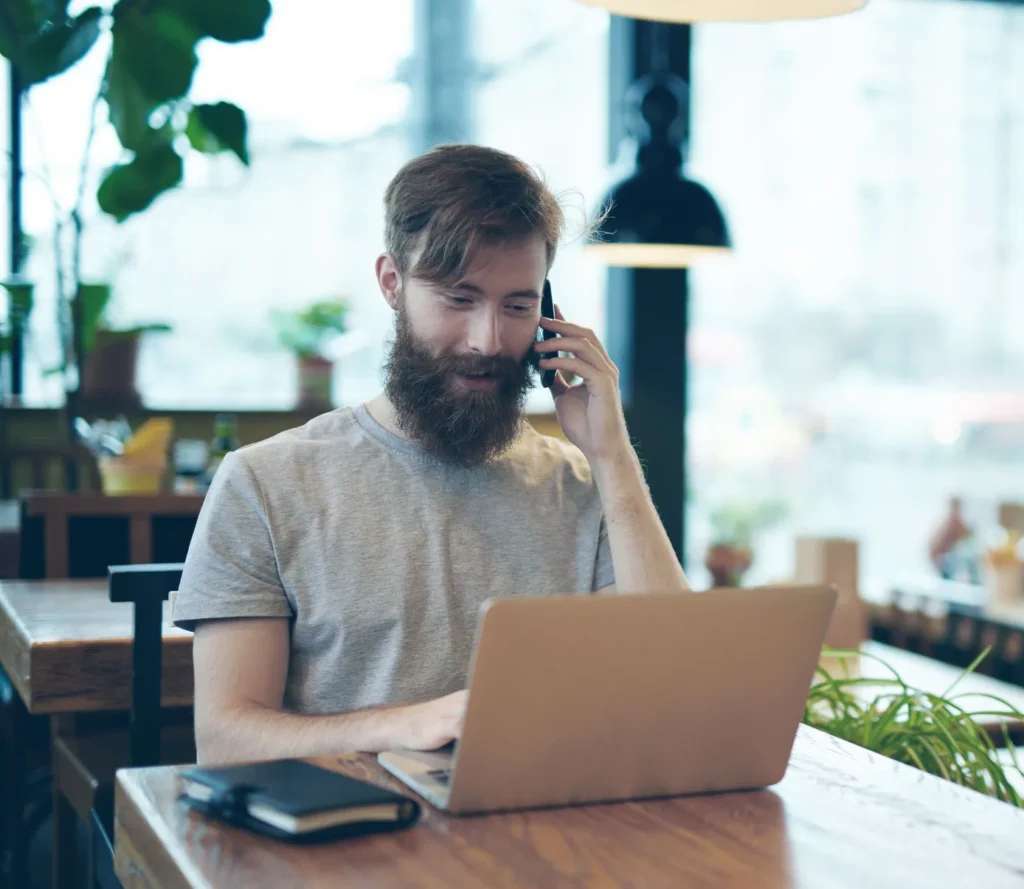 Cheerful business owner discussing project with his client on smartphone while sitting in cozy cafe, waist-up portrait